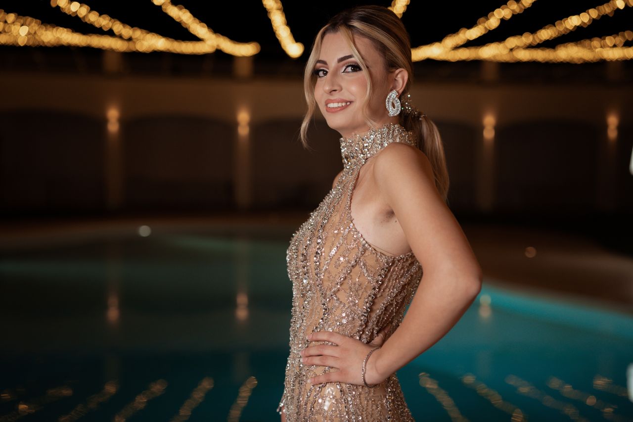 who-we-are Woman in sequined dress poses elegantly by a pool under warm evening lights.